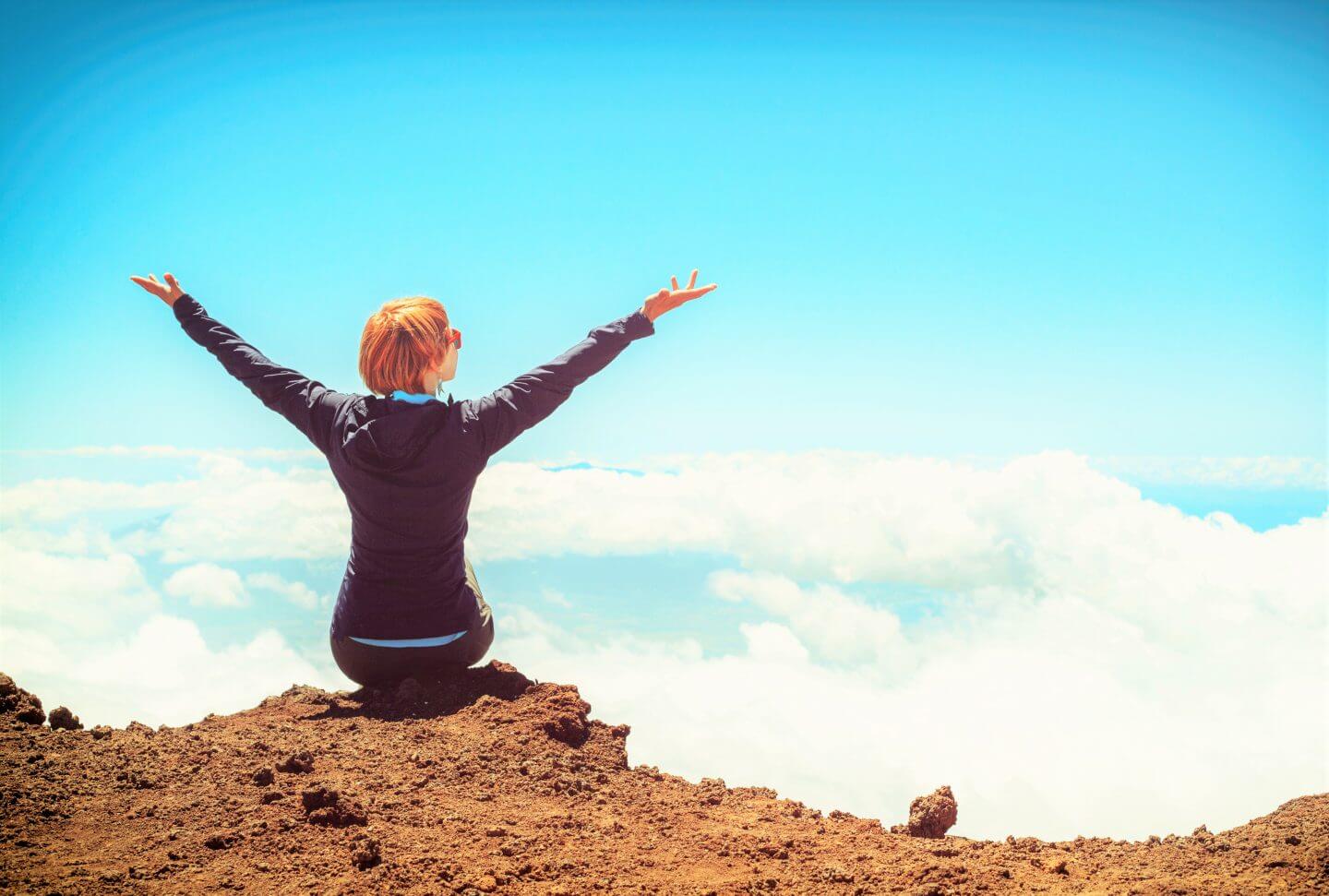 A woman sitting on top of a cliff looking out at the sky with her arms held up towards it.